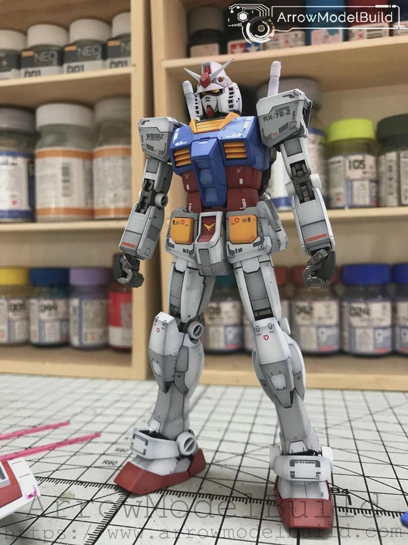 以下が含まれることがあります： 白、青、赤、黄色のガンダムロボットの詳細なモデル。モデルは「ArrowModelBuild」のテキストが見えるグリッド面に立っています。ロボットは複雑な機械的詳細を持っています。