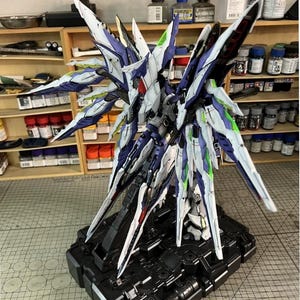 May include: A detailed model of a futuristic robot with large, wing-like structures. The model features a colour scheme of white, blue, and green accents, mounted on a black base. The model is a detailed representation of a science fiction or anime-inspired design.