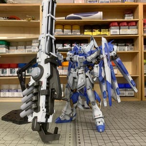 Könnte beinhalten: Detaillierter Modellbausatz eines weißen und blauen Mecha-Roboters mit einer großen Waffe. Der Roboter steht neben seiner Waffe auf einer Schneidematte. Das Bild enthält den Text "ArrowModelBuild" und eine Website-Adresse.