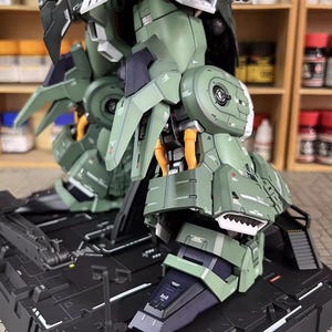 May include: Close-up of a detailed, olive green and black model kit of a mech robot. The model features intricate paneling, articulated joints, and orange accent details. The model is mounted on a black base, showcasing its legs and lower body.