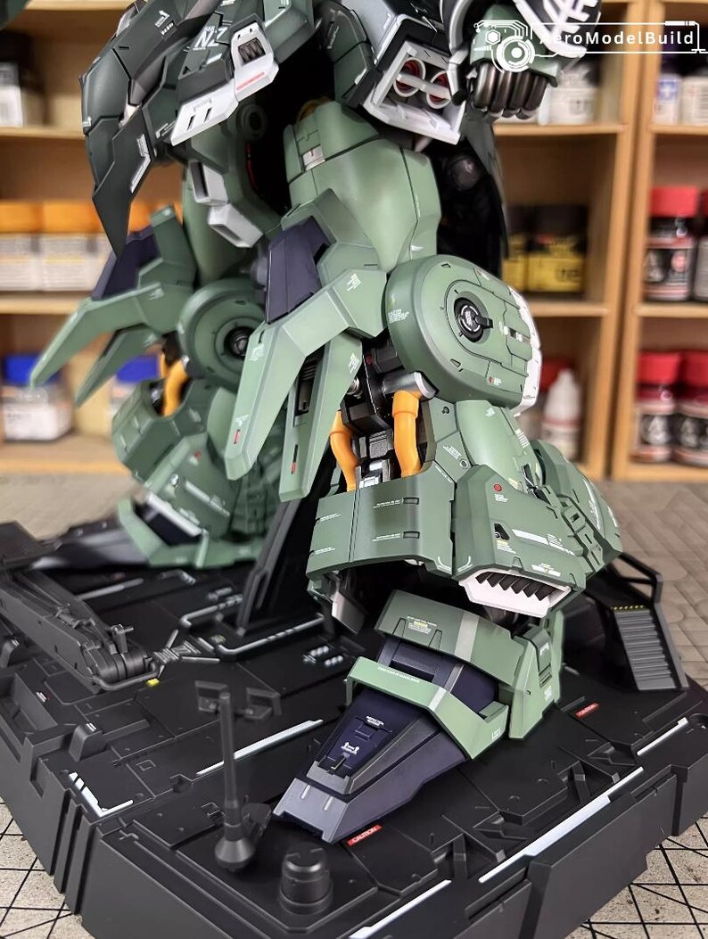 May include: Close-up of a detailed, olive green and black model kit of a mech robot. The model features intricate paneling, articulated joints, and orange accent details. The model is mounted on a black base, showcasing its legs and lower body.