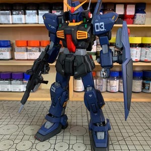 May include: A detailed model of a dark blue and black mecha robot with orange and red accents. The robot has a shield, a rifle, and the number "03" is visible on its body. The background includes shelves with paint bottles and the text "ArrowModelBuild".