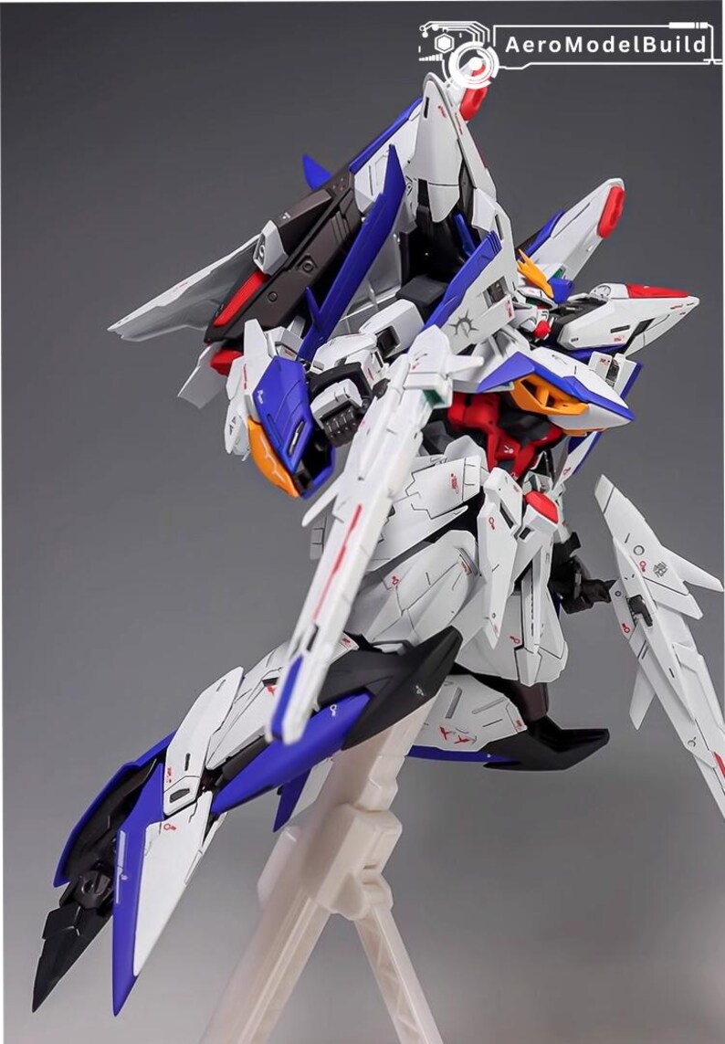 Puede incluir: Kit de modelo detallado de un robot futurista, predominantemente blanco con detalles en azul, rojo y negro. El modelo presenta extremidades articuladas, alas y l&iacute;neas de paneles intrincadas, montado sobre un soporte blanco. El texto "AeroModelBuild" es visible.