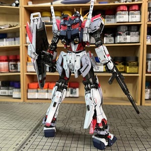 May include: A detailed model of a robot with a white, red, black, and gray color scheme. The robot is standing upright with a shield and a rifle. The model is on a cutting mat with a grid pattern, and the background includes shelves with paint bottles.