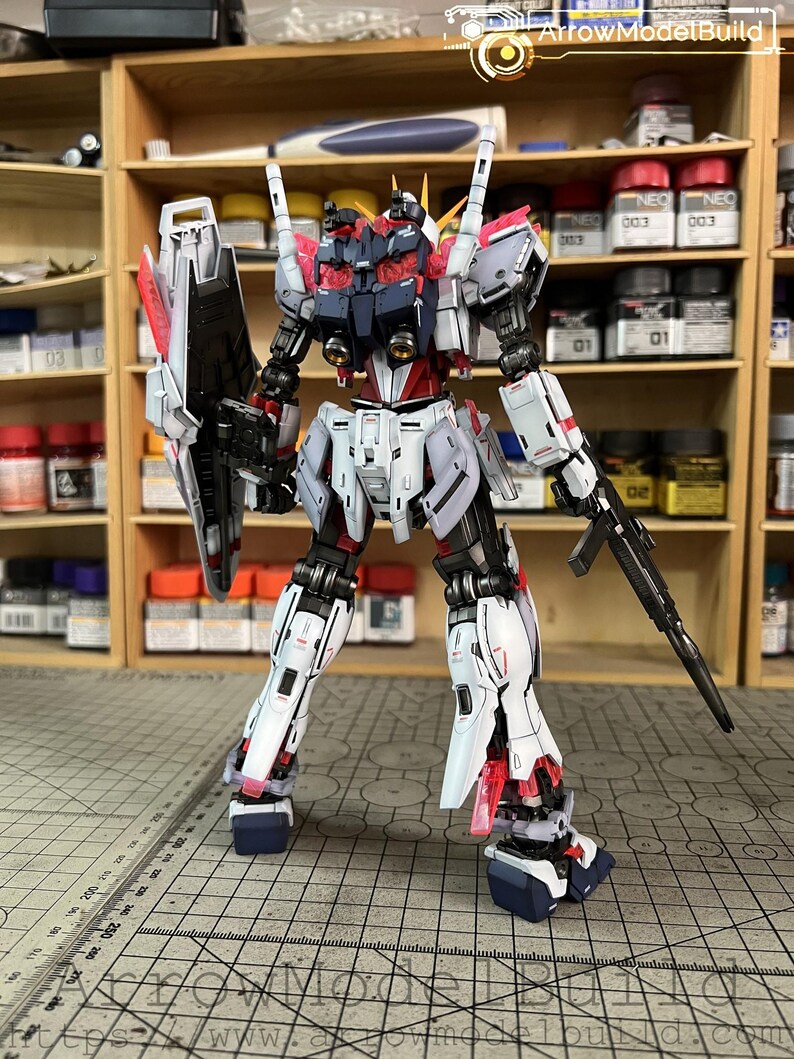 May include: A detailed model of a robot with a white, red, black, and gray color scheme. The robot is standing upright with a shield and a rifle. The model is on a cutting mat with a grid pattern, and the background includes shelves with paint bottles.