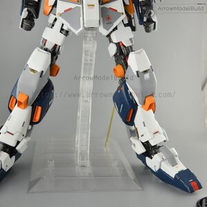 May include: A partially assembled model kit featuring a robotic figure with white, gray, orange, and navy blue components. The model is mounted on a clear plastic stand. The text "ArrowModelBuild" and a website address are visible.