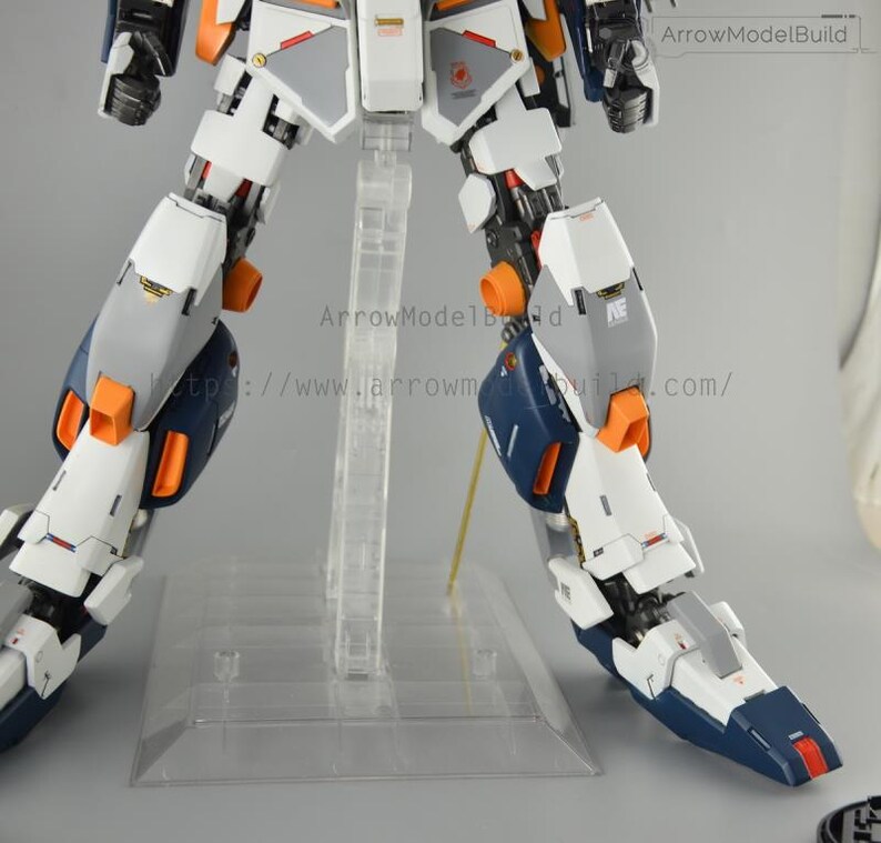 May include: A partially assembled model kit featuring a robotic figure with white, gray, orange, and navy blue components. The model is mounted on a clear plastic stand. The text "ArrowModelBuild" and a website address are visible.