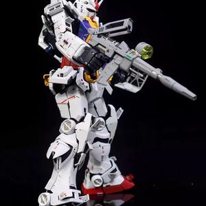 Op de afbeelding: Gedetailleerd model van een witte, rode en grijze robot met een grijs geweer. De robot heeft een gele en blauwe kop met een rode kam. Het model staat op een reflecterend zwart oppervlak.