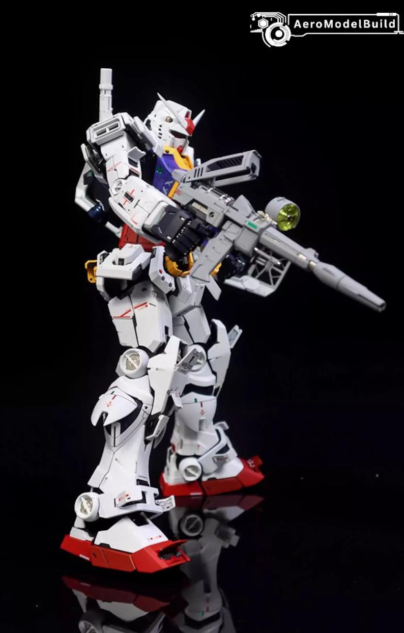 Op de afbeelding: Gedetailleerd model van een witte, rode en grijze robot met een grijs geweer. De robot heeft een gele en blauwe kop met een rode kam. Het model staat op een reflecterend zwart oppervlak.
