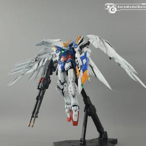 May include: A Wing Gundam Zero model kit featuring large white feathered wings, a blue, white, and red torso, and a twin buster rifle. The figure is mounted on a black display stand and includes the AeroModelBuild logo in the top right corner.
