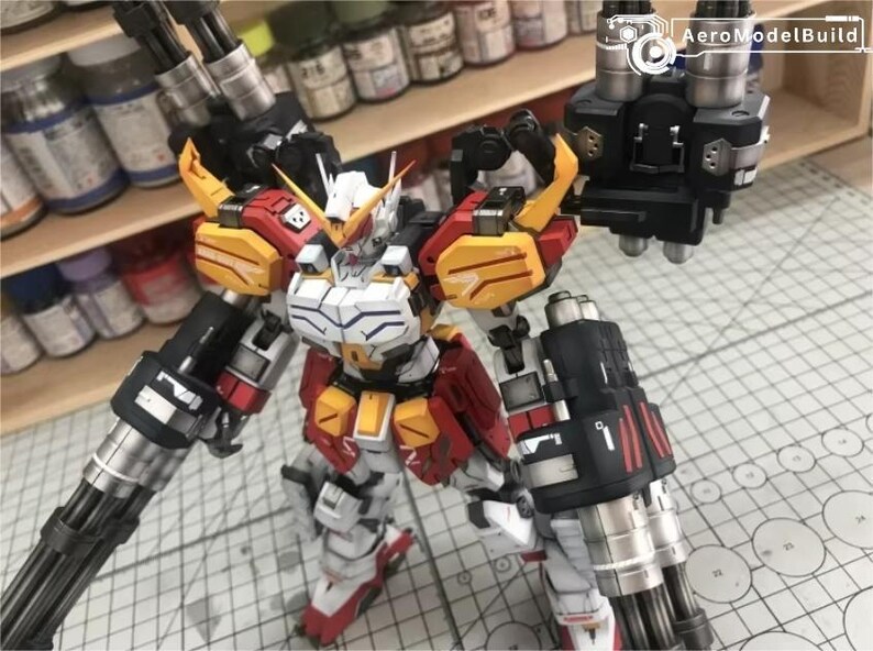 K&ouml;nnte beinhalten: Detailliertes Modell eines Roboters in Wei&szlig;, Rot und Gelb. Der Roboter hat gro&szlig;e Kanonen und steht auf einer Schneidematte. Das AeroModelBuild-Logo ist oben rechts sichtbar.