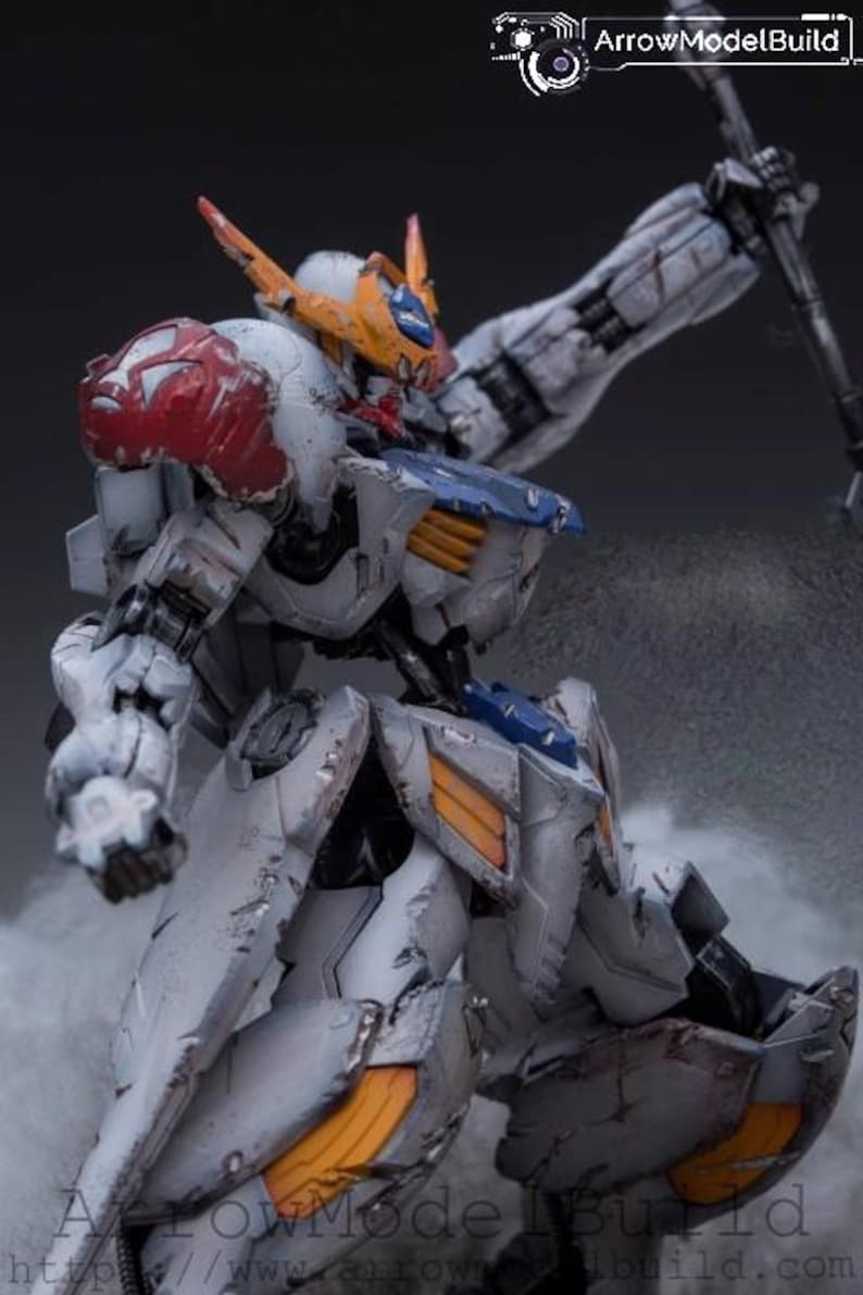 May include: A detailed model of a mecha robot, primarily white with accents of red, blue, and orange. The robot is posed dynamically, with a weapon raised. The model has a weathered appearance, suggesting battle damage. The text "ArrowModelBuild" is visible.