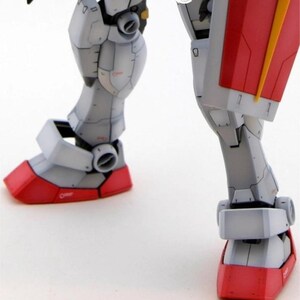 May include: Close-up of a detailed model robot's legs. The robot's legs are primarily gray with red accents on the feet and a yellow detail. The model has visible panel lines and mechanical details, suggesting a complex design.