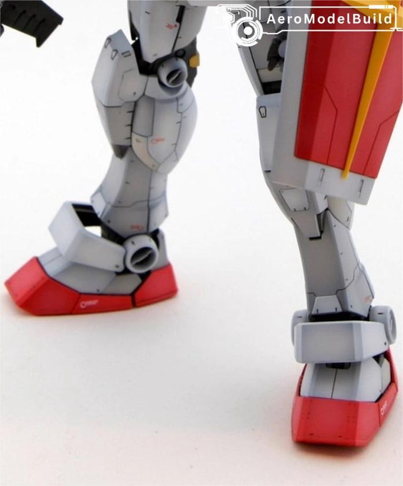 May include: Close-up of a detailed model robot's legs. The robot's legs are primarily gray with red accents on the feet and a yellow detail. The model has visible panel lines and mechanical details, suggesting a complex design.
