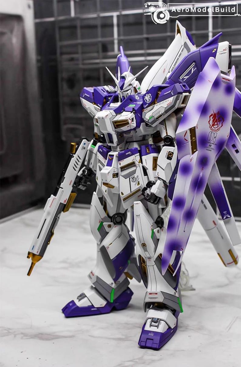 May include: A detailed model of a Gundam robot, primarily white with purple and gold accents. It features a large shield, a long rifle, and intricate mechanical detailing. The model is posed on a neutral surface.