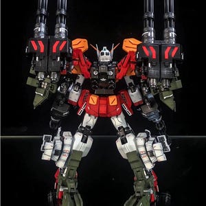 K&ouml;nnte beinhalten: Eine mehrfarbige Roboter-Actionfigur mit zwei gro&szlig;en schwarzen Gatling-Kanonen an den Schultern, rot-wei&szlig;er Panzerung und olivgr&uuml;nen Beinkomponenten. Der Text AeroModelBuild ist in der oberen rechten Ecke des Bildes zu sehen.