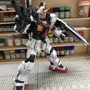 Op de afbeelding: Gedetailleerd model van een Gundam-robot, voornamelijk wit met zwarte, rode en gele accenten. De robot poseert met een wapen en schild, tegen een achtergrond van verfbenodigdheden en planken.
