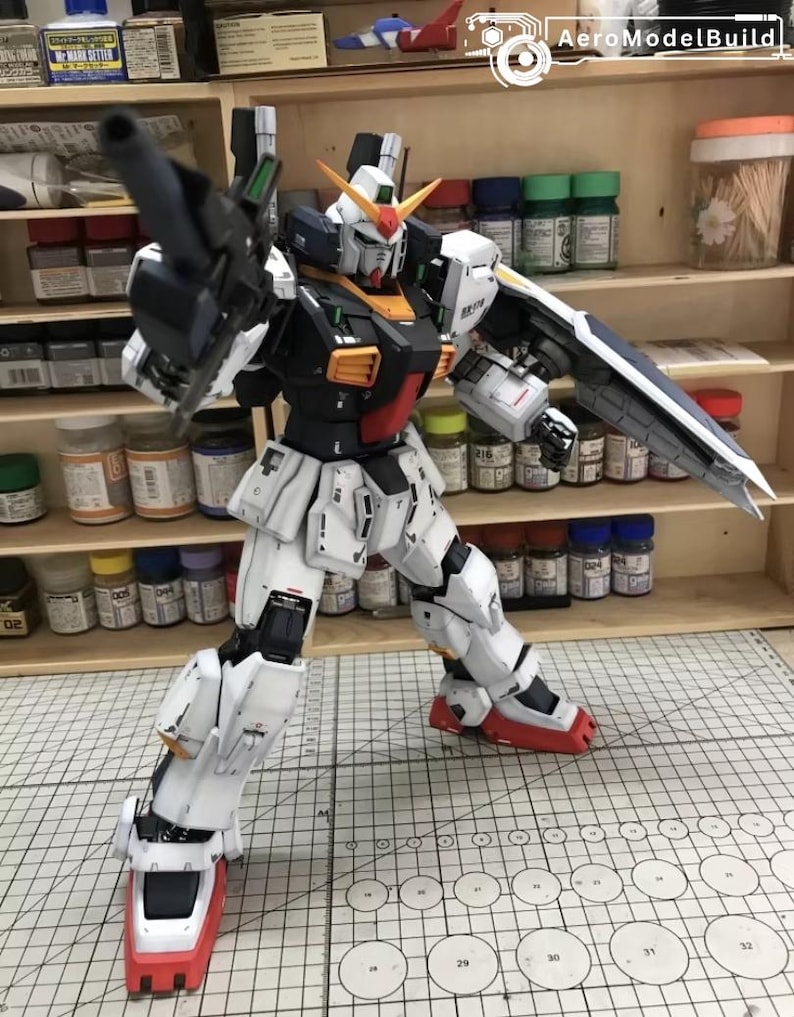 Op de afbeelding: Gedetailleerd model van een Gundam-robot, voornamelijk wit met zwarte, rode en gele accenten. De robot poseert met een wapen en schild, tegen een achtergrond van verfbenodigdheden en planken.
