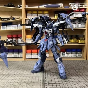 May include: A detailed model of a blue and gray mech robot with articulated limbs and weapons. The robot has a complex design with multiple panels, wings, and a detailed head. The background shows shelves filled with model-building supplies.