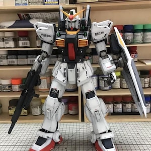Op de afbeelding: Gedetailleerd model van een witte, zwarte en rode robot met een groot schild en geweer. De robot heeft meerdere panelen en gewrichten, met gele en oranje accenten. Het staat voor een plank met verfpotten.