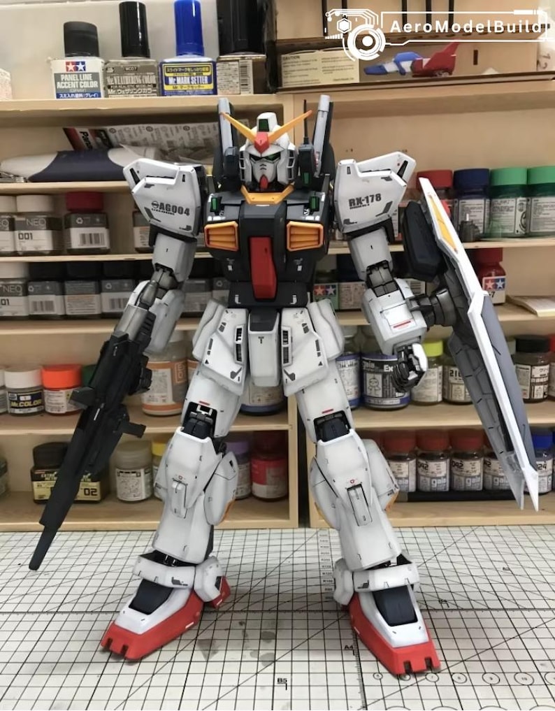 Op de afbeelding: Gedetailleerd model van een witte, zwarte en rode robot met een groot schild en geweer. De robot heeft meerdere panelen en gewrichten, met gele en oranje accenten. Het staat voor een plank met verfpotten.