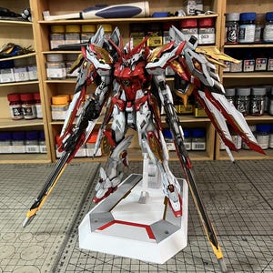May include: A detailed model of a red, white, and gold mecha robot with large wings and two long swords. The robot is standing on a white base with red accents. The background includes shelves with paint bottles and tools.