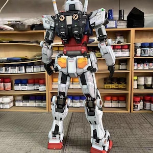 May include: A detailed, assembled model of a Gundam robot, primarily white with red, blue, and black accents. The model features intricate panel lines and mechanical details, with visible joints and a complex design. The robot stands on a cutting mat.