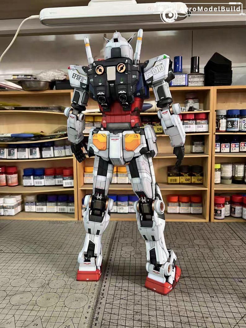 May include: A detailed, assembled model of a Gundam robot, primarily white with red, blue, and black accents. The model features intricate panel lines and mechanical details, with visible joints and a complex design. The robot stands on a cutting mat.