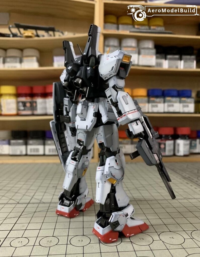Pode incluir: Um modelo detalhado de um rob&ocirc; mecha branco, cinza, preto e vermelho com uma arma e um escudo. O modelo tem detalhes intrincados e est&aacute; posicionado em uma superf&iacute;cie de grade. O texto "AeroModelBuild" &eacute; vis&iacute;vel.
