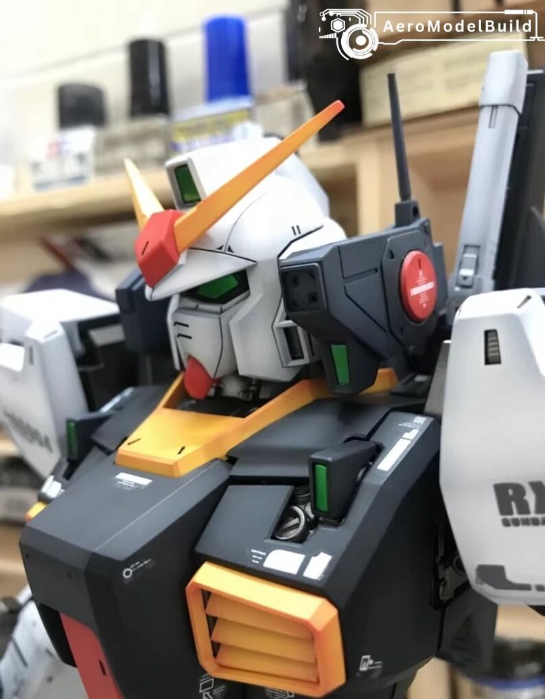 Op de afbeelding: Een gedetailleerd model van een Gundam-robot, met een witte kop en rode en gele accenten. Het model heeft zwarte en gele bepantsering, met groene en rode details. De tekst "AeroModelBuild" is zichtbaar in de rechterbovenhoek.