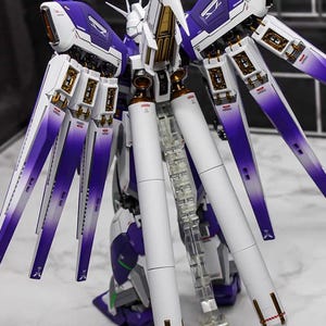 May include: A detailed model kit of a futuristic robot, featuring a color scheme of white, purple, and gold. The model has multiple articulated wings and a clear stand. The text "AeroModelBuild" is visible.