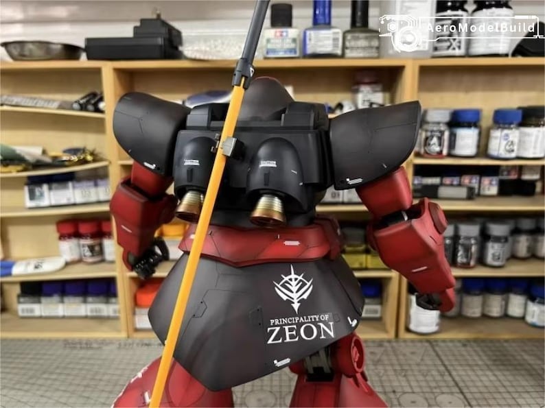 May include: A detailed model of a red and black mecha robot with an orange pole. The robot has a black torso with the text "PRINCIPALITY OF ZEON". The background shows shelves with paint bottles.