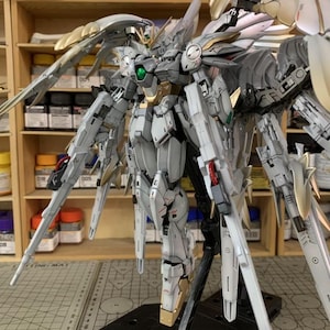 May include: A detailed, assembled Gundam model kit in shades of white, gray, and gold. The model features intricate mechanical details, large wings, and multiple weapons. The model is mounted on a black stand, with a workshop background.