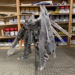 May include: A transparent plastic model robot with articulated limbs and large wings. The model is partially assembled and stands on a grid-patterned surface. The background includes shelves with paint bottles and tools.