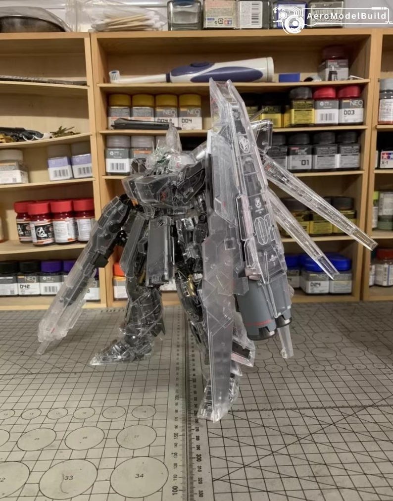 May include: A transparent plastic model robot with articulated limbs and large wings. The model is partially assembled and stands on a grid-patterned surface. The background includes shelves with paint bottles and tools.