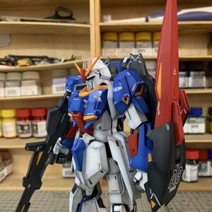 May include: A detailed model robot, predominantly blue, white, and red, with a black rifle and a large red shield. The robot features intricate mechanical details and is set against a backdrop of wooden shelves.