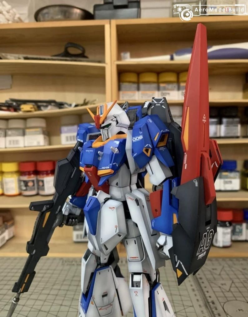 May include: A detailed model robot, predominantly blue, white, and red, with a black rifle and a large red shield. The robot features intricate mechanical details and is set against a backdrop of wooden shelves.