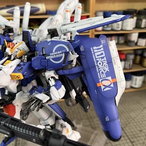 May include: A detailed model of a Gundam robot, mainly blue and white, with intricate mechanical details. The model features text that reads "110 TASK FORCE" and "EARTH FEDERATION SPACE FORCE". The model is on display with a shelf of paint bottles in the background.