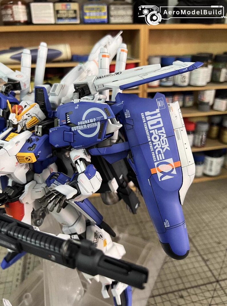 May include: A detailed model of a Gundam robot, mainly blue and white, with intricate mechanical details. The model features text that reads "110 TASK FORCE" and "EARTH FEDERATION SPACE FORCE". The model is on display with a shelf of paint bottles in the background.