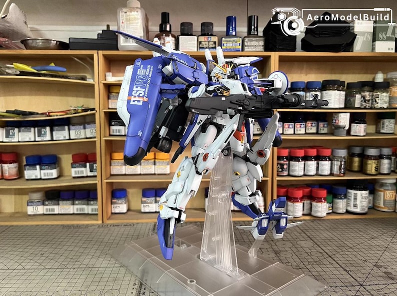 May include: A detailed model robot, mainly white and blue, holding a large weapon. The model is displayed on a clear stand. The text "EFSF PG" is visible on its back. Shelves with bottles and containers are in the background.