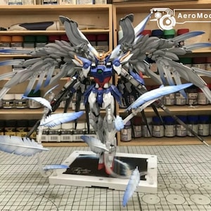 May include: A Wing Gundam Zero model kit featuring a blue, white, and grey color scheme with large articulated wings. The figure is displayed on a white base with floating feather accessories and includes the text AeroModelBuild.