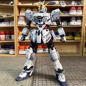 May include: A detailed, articulated white and gray Gundam model kit with black accents and red detailing. The model has a complex design with multiple panels and joints, and yellow head crests. The model is posed in a standing position.