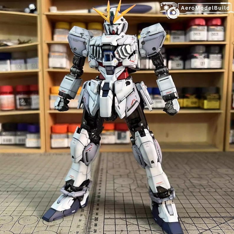 May include: A detailed, articulated white and gray Gundam model kit with black accents and red detailing. The model has a complex design with multiple panels and joints, and yellow head crests. The model is posed in a standing position.