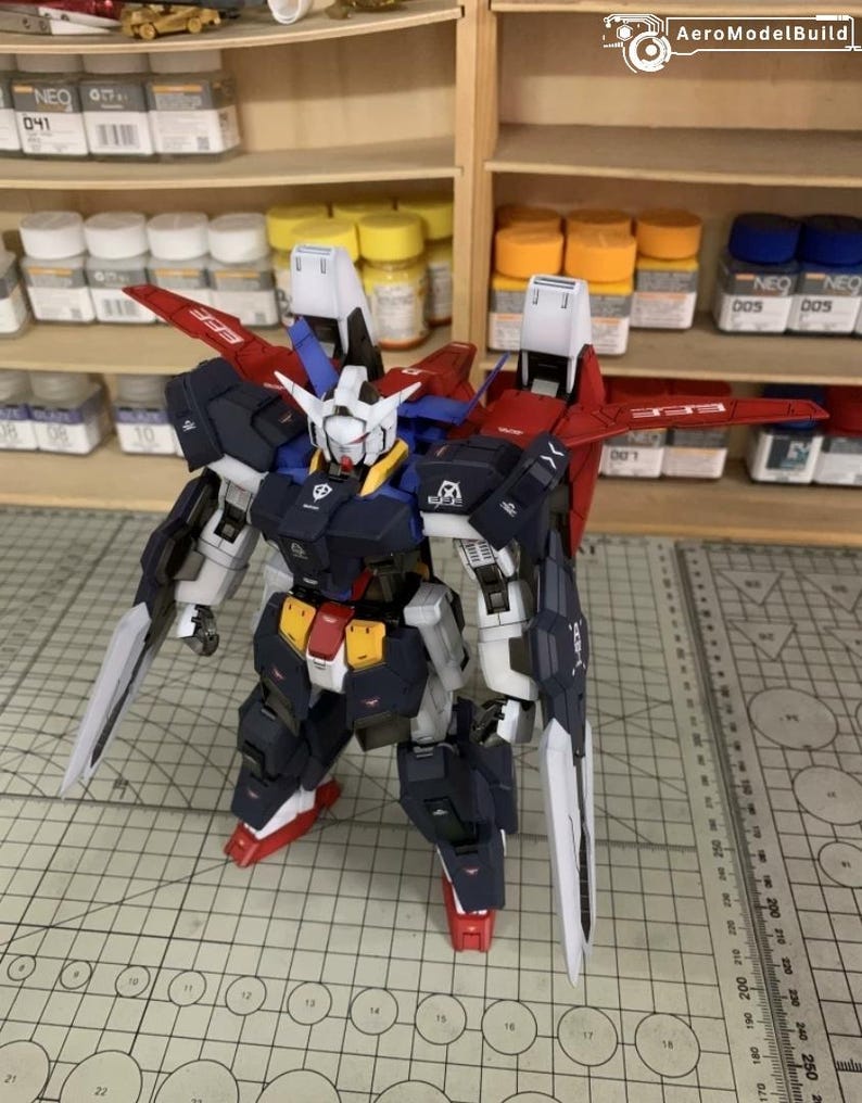 Puede incluir: Kit de modelo detallado de un robot mecha, con una combinaci&oacute;n de colores azul marino, blanco, rojo y amarillo. El robot tiene grandes alas y est&aacute; equipado con dos espadas largas y blancas. El modelo est&aacute; posado sobre una esterilla de corte.