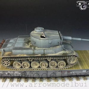 Puede incluir: Modelo detallado de un tanque gris sobre un vagón de plataforma. El tanque tiene un cañón largo y está desgastado con suciedad y mugre. La plataforma está hecha de tablones de madera y el vagón se asienta sobre una vía. El modelo es una pieza de exhibición.