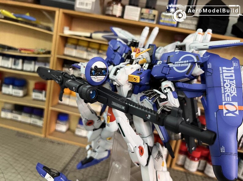 May include: A detailed model robot in blue, white, and yellow, holding a black rifle. The robot features a shield with the text "110 TASK FORCE" and "EARTH FEDERATION SPACE FORCE". The model is set against a background of shelves.