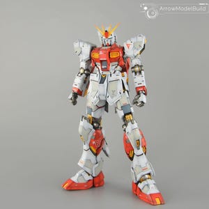 May include: A detailed model of a Gundam robot, predominantly white with red and yellow highlights. The robot features intricate mechanical detailing, including articulated joints and panels. The model is standing upright against a neutral grey background.