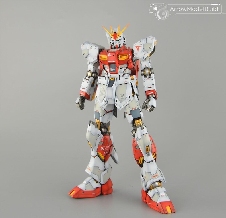 May include: A detailed model of a Gundam robot, predominantly white with red and yellow highlights. The robot features intricate mechanical detailing, including articulated joints and panels. The model is standing upright against a neutral grey background.