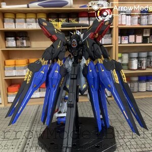 Puede incluir: Un detallado kit de modelo de un mech con detalles en negro, azul, rojo y dorado. El modelo tiene alas grandes y est&aacute; posado sobre un soporte negro. El fondo muestra estantes con varias botellas y recipientes.