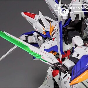 Puede incluir: Modelo detallado de un robot mecha blanco, azul, rojo y amarillo con una espada de energ&iacute;a verde. El robot tiene dise&ntilde;os intrincados y est&aacute; posado din&aacute;micamente. El texto "AeroModelBuild" es visible en la esquina superior derecha.